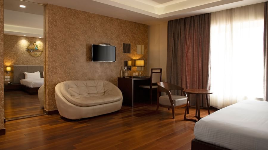 A spacious room featuring a large bed, a sofa, a flat-screen TV mounted on the wall, and wooden flooring at The Hamuse Hotel, Kodaikanal.