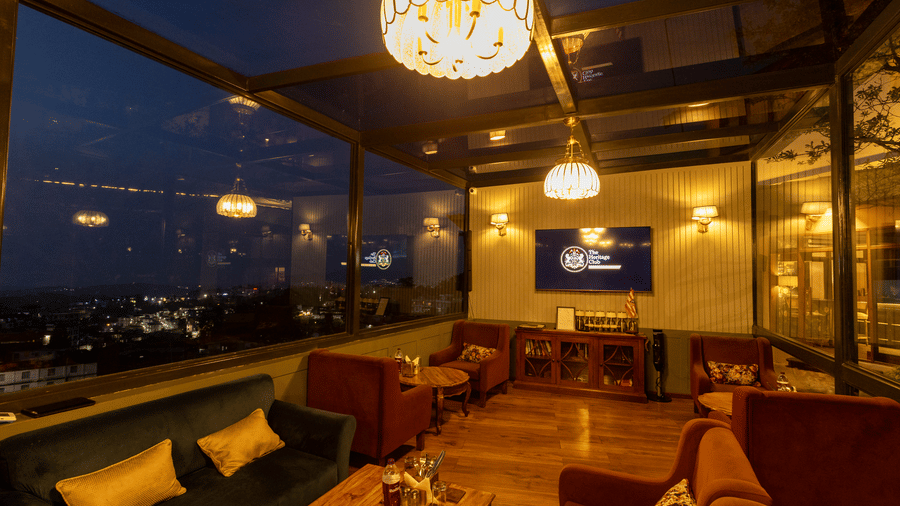 A dimly lit rooftop lounge at night featuring low sofa seating, wooden floors, and large glass windows overlooking city lights. | The Heritage Club, Tripura, Castle, Shillong