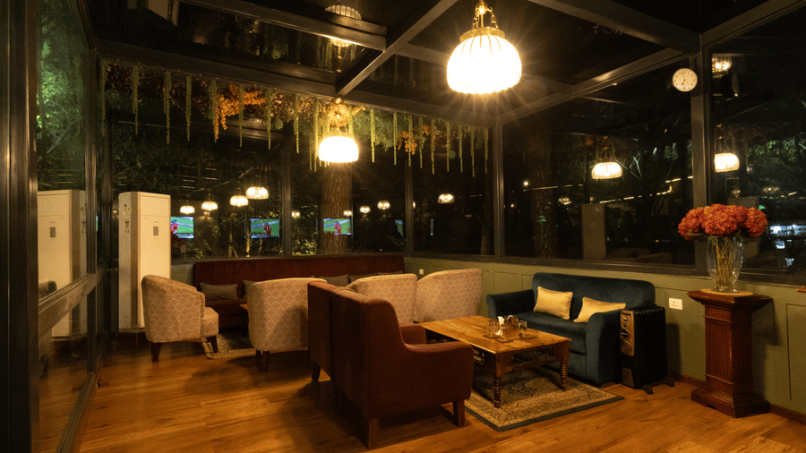 An indoor lounge area featuring a large plush sofa arranged around a low table. The space is illuminated by a spherical overhead light and bordered by large windows overlooking an outdoor area. | The Heritage Club, Tripura, Castle, Shillong