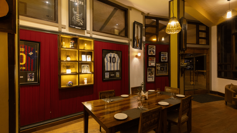 An indoor dining area featuring dark wooden tables and chairs set against deep red walls adorned with framed sports jerseys. | The Heritage Club, Tripura, Castle, Shillong