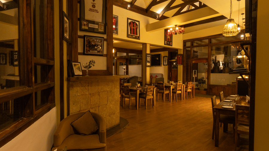 A restaurant with polished flooring and numerous tables. Built-in wooden cabinetry runs along the left wall, and multiple light fixtures hang from the ceiling. | The Heritage Club, Tripura, Castle, Shillong