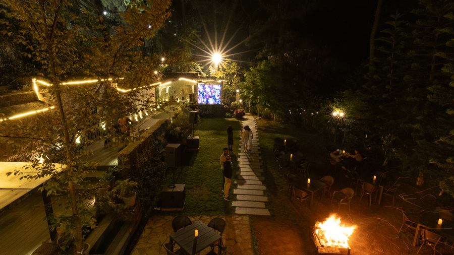 An illuminated lawn showcasing chairs, and space set with campfire, with people, and trees. | The Heritage Club