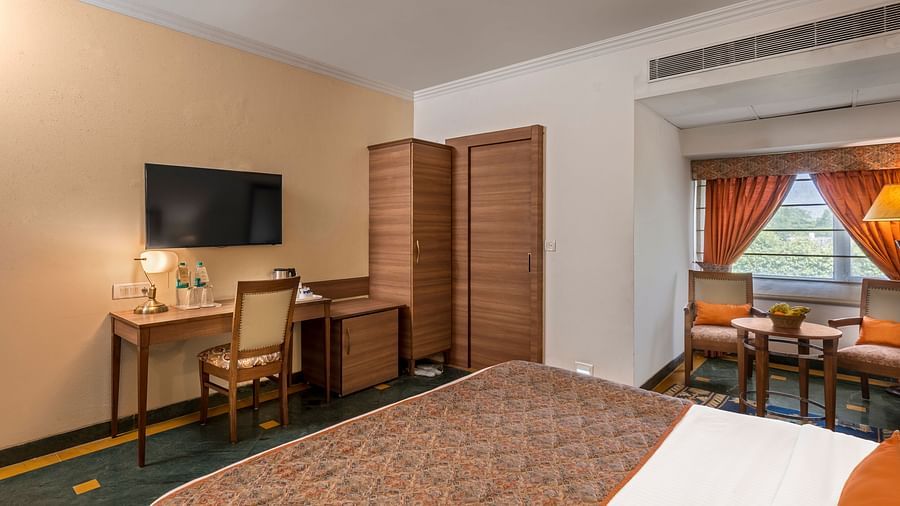 side view of a bedroom with a bed facing a television, below which is a workdesk and chair and a wardrobe beside it - The Manor, Kashipur