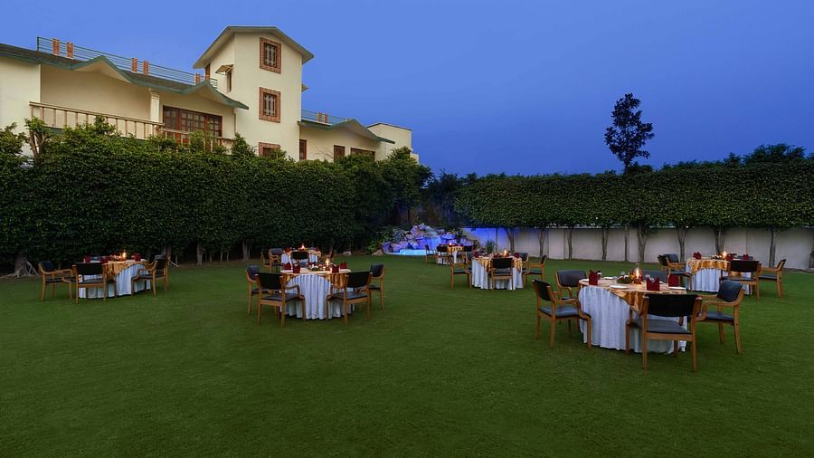 An open lawn with tables and chairs arranged in a circle, each adorned with decorative settings and a small lamp, featuring a lit-up fountain in the corner