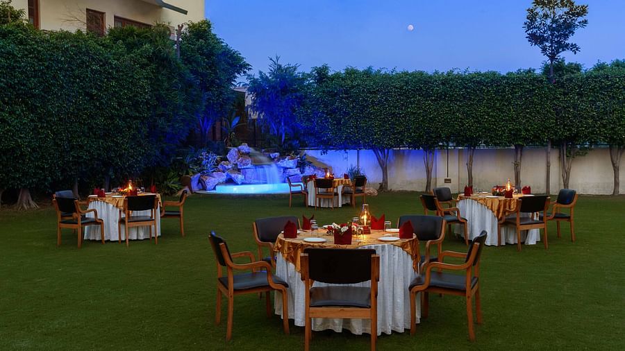 An open lawn with tables and chairs arranged in a circle, each adorned with decorative settings and a small lamp, featuring a lit-up fountain in the corner