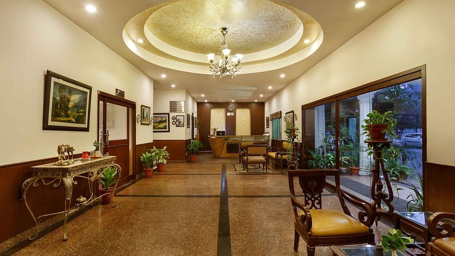 A corridor adorned with decorative frames, featuring high tables topped with flower vases and indoor plants, along with a few chairs arranged along the sides