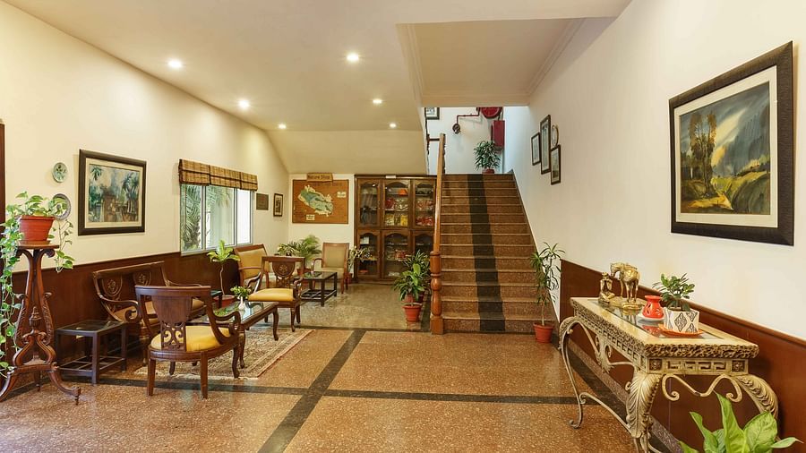 A corridor adorned with photo frames on the walls, high tables with flower pots on top, chairs along the side, and stairs leading to the next floor