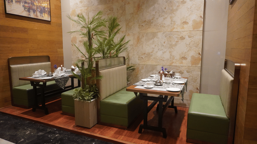 Restaurant seating, wooden table set with napkins and glassware. Decorative plants divide the booths, under warm lighting at the Meriton. 