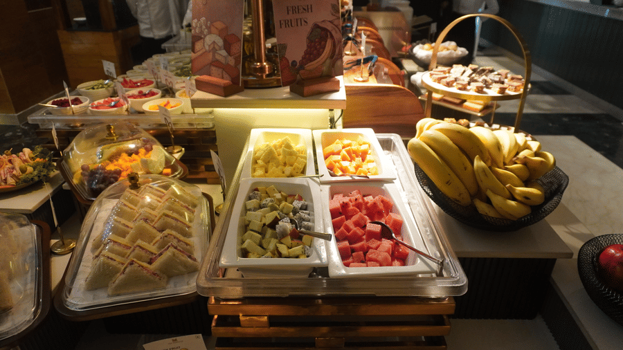 A variety of fresh, colorful fruits and prepared food items are displayed in white rectangular trays and bowls on a buffet table   at the Meriton.