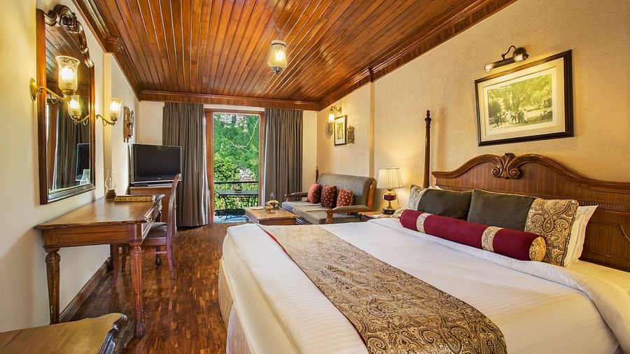 An image of a hill view room in naini retreat, nainital, with a king-size bed, wooden furniture, and a bay window overlooking a garden. The room features a ceiling fan, a television, and a small sitting area.