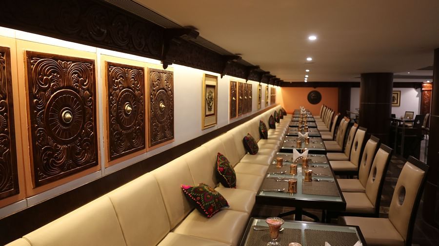 A cosy restaurant interior featuring an extended cream sofa, ornate wooden panels on the wall, and neatly set tables for dining at The President Hotel, Jayanagar.