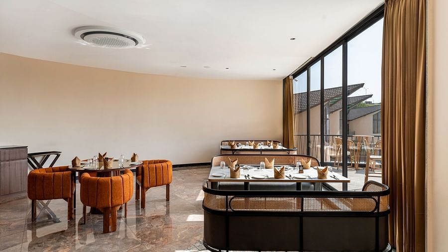 Fine Dine with warm lighting, featuring wooden tables and chairs, a comfortable sofa, and large windows offering a view of the outside at The Qualia Resort Club & Brewery, Udaipur.