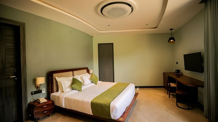 Valley View Room furnished with a king size bed dressed in white linens and a green accent runner, complemented by a wooden side table and a television mounted on the wall at The Qualia Resort Club & Brewery, Udaipur.