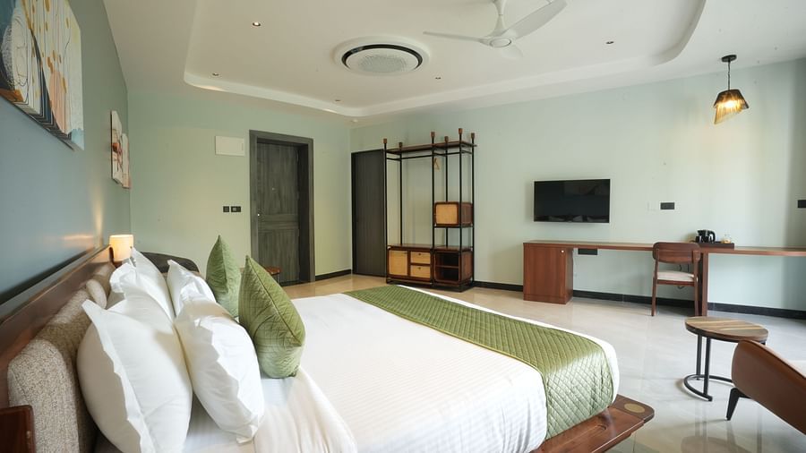 Club Suite with Valley View Sitout featuring a king size bed with white linens and green accents, several pillows, and a wooden headboard at The Qualia Resort Club & Brewery, Udaipur.