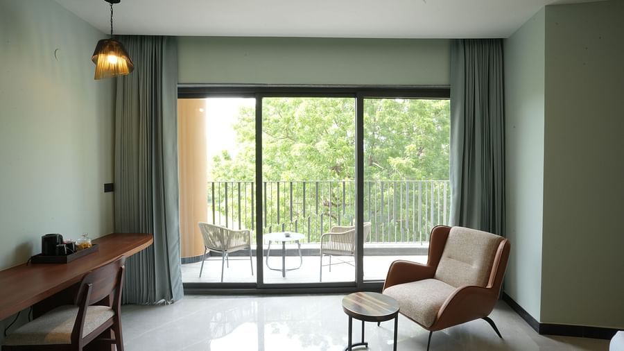 Club Suite with Valley View Sitout depicts a contemporary living space featuring a desk, chair, lounge chair, and a large window offering a view of a balcony and greenery at The Qualia Resort Club & Brewery, Udaipur.