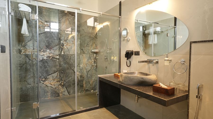 A modern bathroom of Club Suite with Valley View Sitout with a glass-enclosed shower, a unique-shaped mirror, and a sleek vanity area at The Qualia Resort Club & Brewery, Udaipur.
