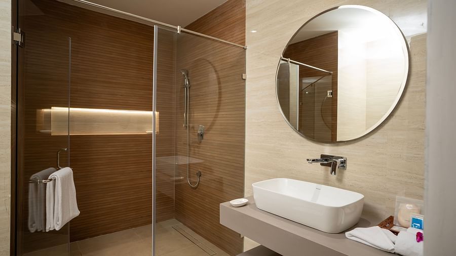 The bathroom featuring a wash basin along with a round mirror above it and a shower area separated by tall glass doors at The Shore, Trishvam.