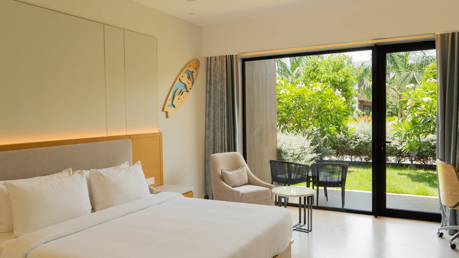 Bright bedroom with a king size bed, seating area and glass doors opening to a balcony with garden views at The Shore Trishvam, Pondicherry.