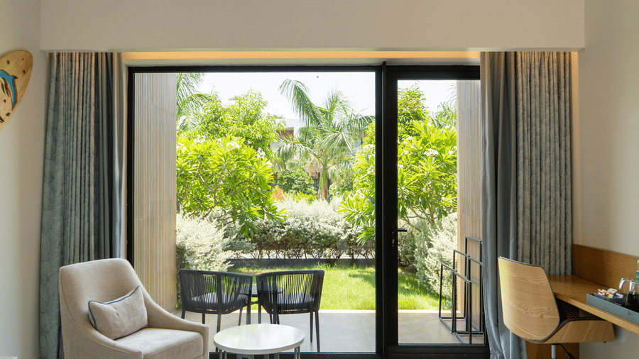 Bright Garden Courtyard bedroom at The Shore Trishvam, Pondicherry, with king bed, surfboard wall art & glass doors overlooking a green garden.