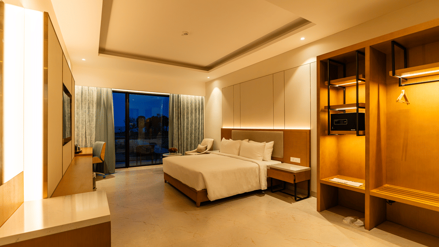 modern hotel room interior at The Shore Trishvam, Pondicherry, featuring large bed, bedside lamps, wardrobe, and glass doors with sheer curtains.