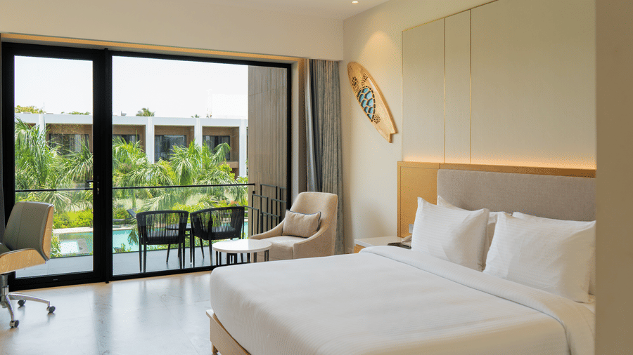 Pool View Bliss room at The Shore Trishvam, Pondicherry, with king bed, surfboard wall art & balcony overlooking the pool and tropical palms.