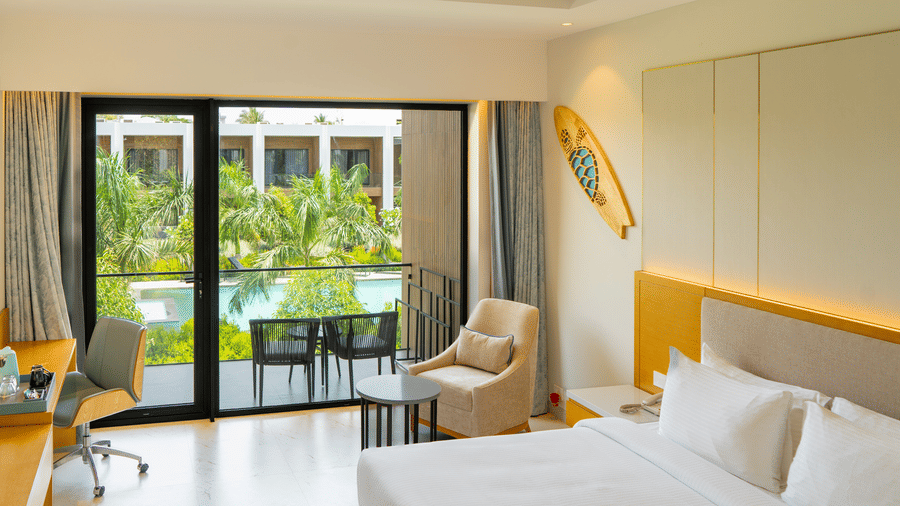 Sunlit Pool View Bliss room at The Shore Trishvam, Pondicherry, featuring a king bed, lounge chair & balcony with pool and palm tree views.
