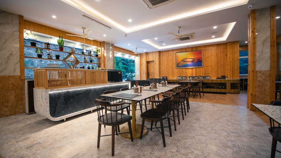 A restaurant at The Tattva Devaprayag with a long wooden table with black chairs and a bar.