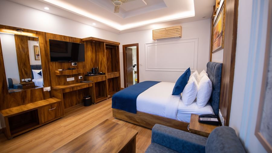 A Deluxe Room at The Tattva Devaprayag with a bed featuring blue blankets and two pillows, a desk, a television, and a sofa.