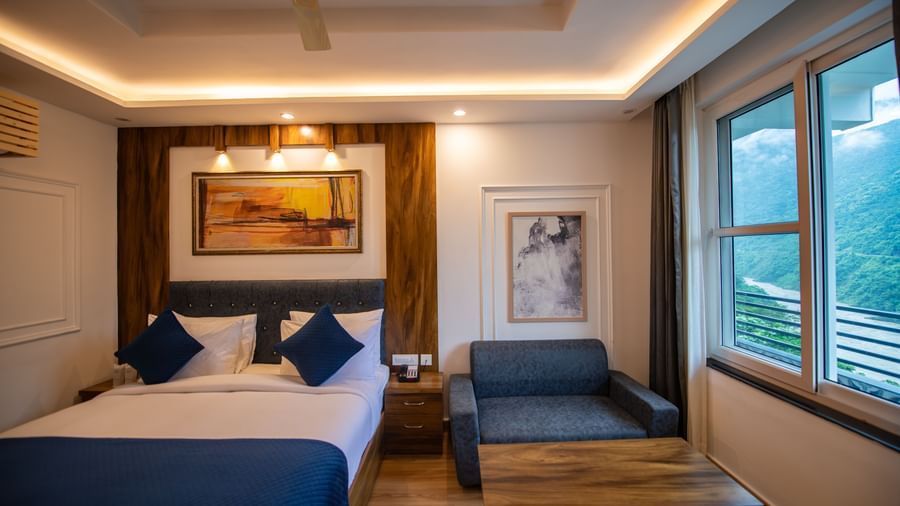 A Deluxe Room at The Tattva Devaprayag with a bed featuring blue blankets and two pillows, two framed paintings on the wall and a sofa.