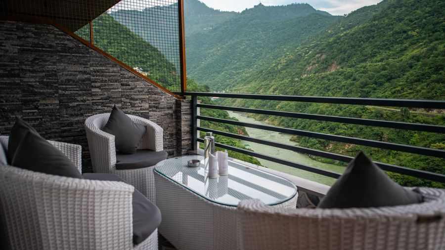 A balcony in the executive room at The Tattva Devaprayag featuring two white chairs and a table with a view of the mountains and the river.