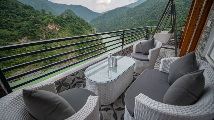 A wide view of mountains and river flowing, a seen from the balcony of The Tattva Devaprayag featuring two grey chairs, a table and a swing.