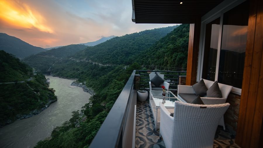  A wide view of the mountains and a river as seen from the balcony of The Tattva Devaprayag featuring a white couch and a table during sunset.