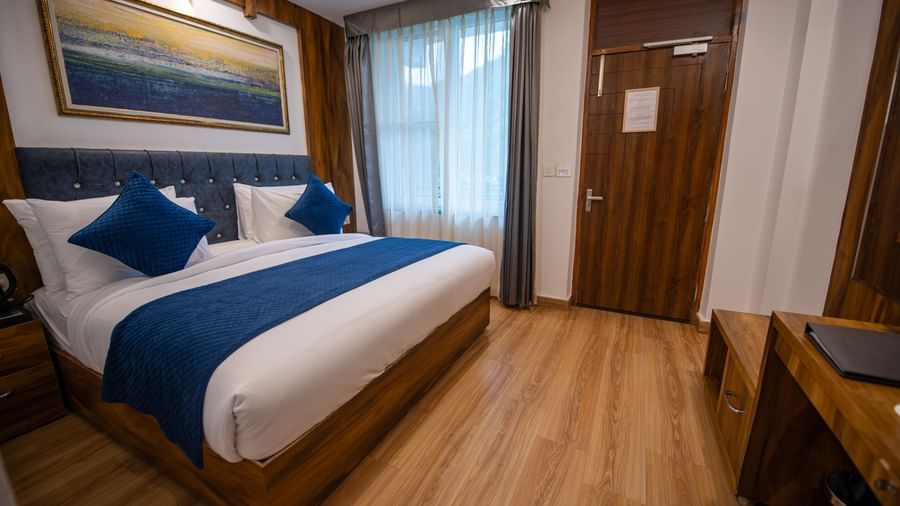A Standard Room at The Tattva Devaprayag with a bed featuring blue blankets and two pillows, a wooden cabinet, and a framed painting on the wall.