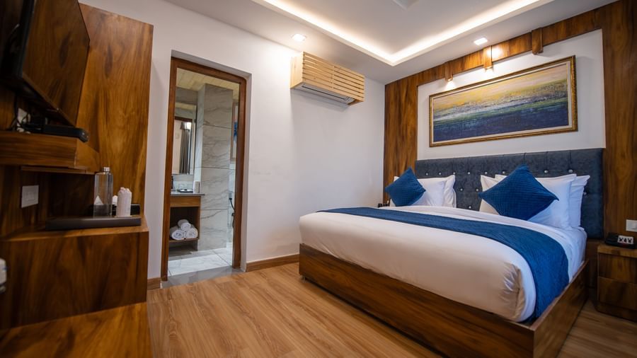 A standard room at The Tattva Devaprayag with a bed featuring blue blankets and two pillows and an open bathroom door.