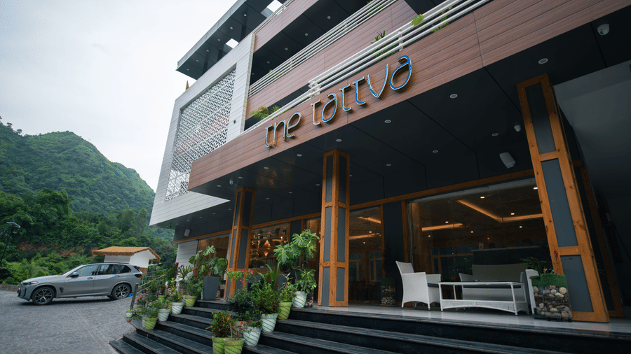 Facade of The Tattva, Devaprayag, featuring steps leading to the entrance, a car parks next to the hotel, trees in the background and cloudy sky above.