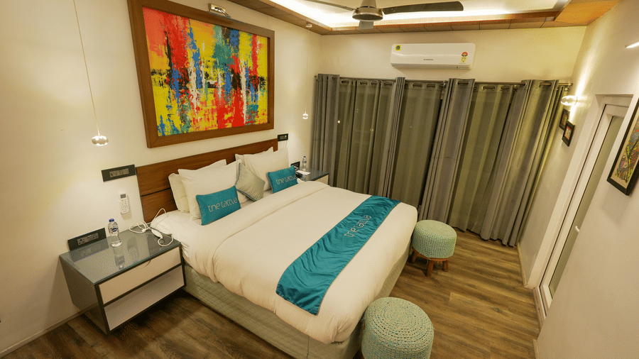 Bedroom of Pent House at The Tattva, Joshimath with a large bed, teal accents, abstract wall art, wooden flooring, grey curtains, and ambient ceiling lighting.