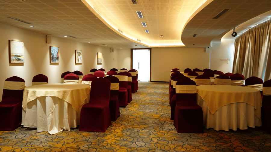 A banquet hall at Tiara by MPS features round tables draped in cream cloths and surrounded by chairs with maroon covers under a wavy recessed ceiling.
