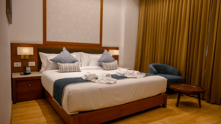 A side perspective of a king size bed flanked by a wooden nightstand and a blue armchair with a circular coffee table at Tiara by MPS.