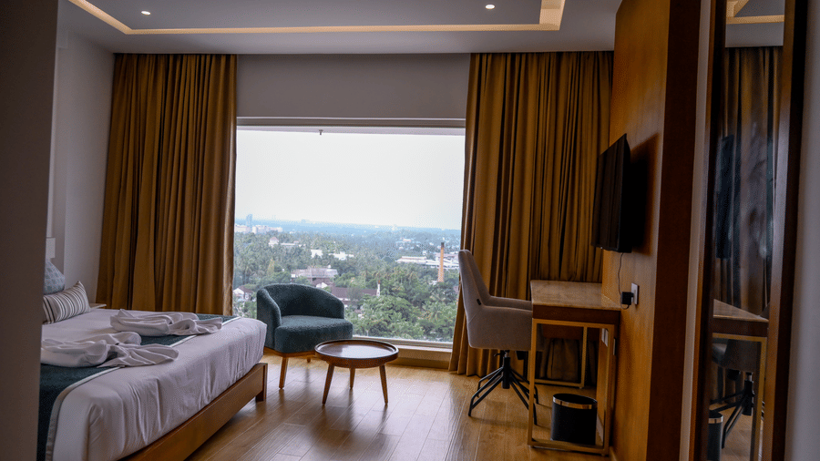 A bedroom view focused on a wide window showing lush green trees outside, complete with an armchair and a study desk setup at Tiara by MPS.