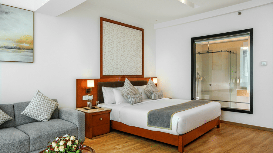 A bedroom showcasing a king size bed alongside a grey sofa and a glass partition looking directly into an attached bathroom at Tiara by MPS.