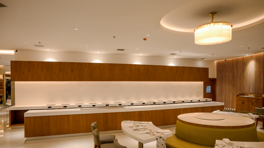 A long buffet counter with a wooden finish and recessed lighting stretches across a dining area with round tables and curved booth seating at Tiara by MPS.