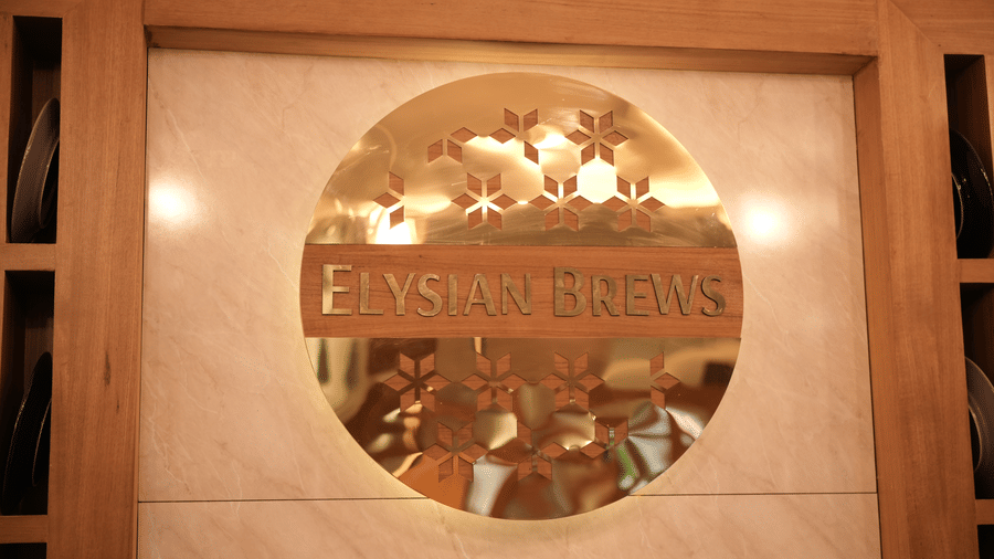 A circular gold reflective sign displays the text Elysian Brews inside a structured rectangular wooden wall frame at Tiara by MPS.
