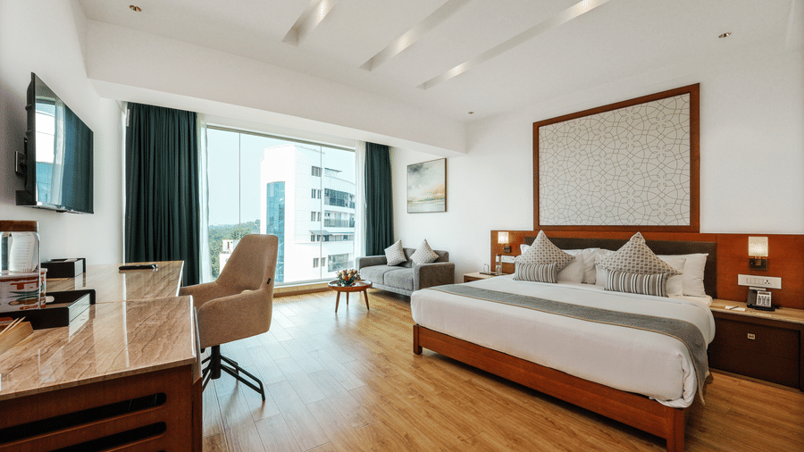 A spacious bedroom featuring a king size bed, a study desk with an armchair, and a large window looking out towards buildings at Tiara by MPS.