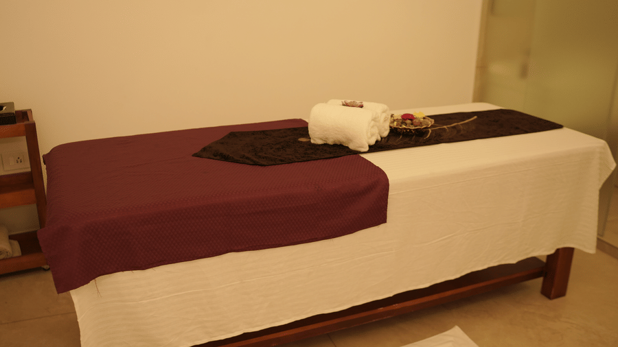 A wooden massage table draped with white and purple sheets stands in a brightly lit spa treatment room with tiled flooring at Tiara by MPS.