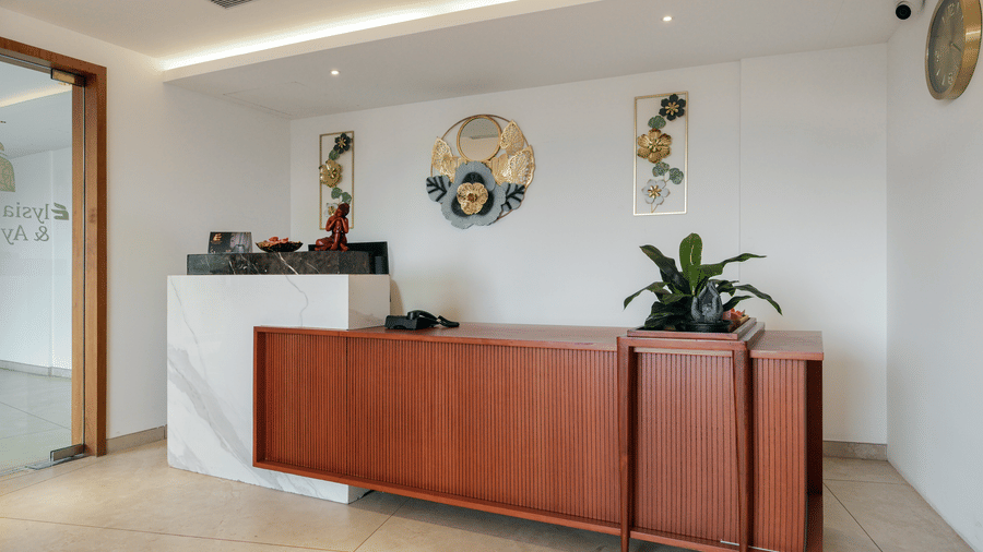 A reception area at Tiara by MPS features a long wooden desk with a white marble counter section and floral decorations mounted on the back wall.