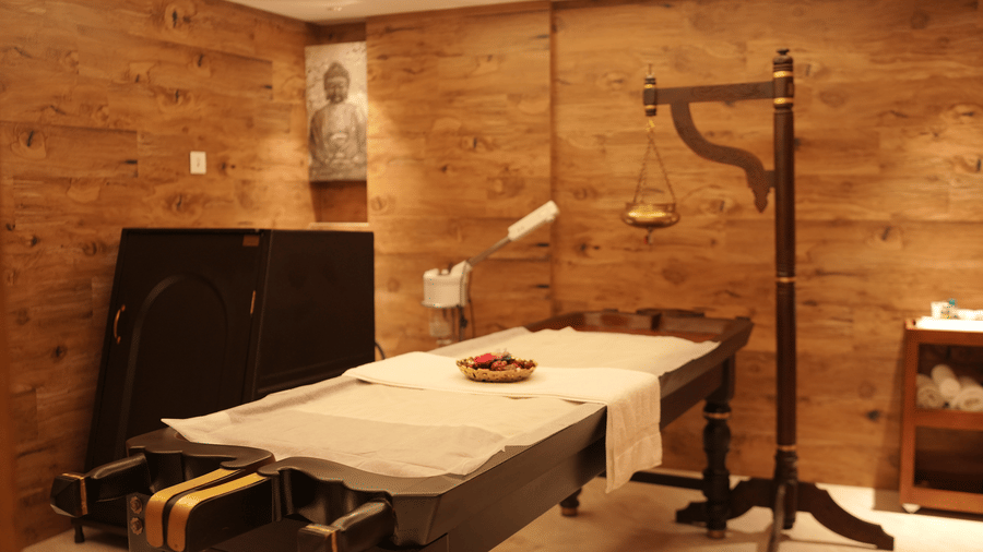 An authentic wooden Ayurvedic massage table with a shirodhara stand is positioned in a wood-panelled therapy room featuring a Buddha portrait at Tiara by MPS.