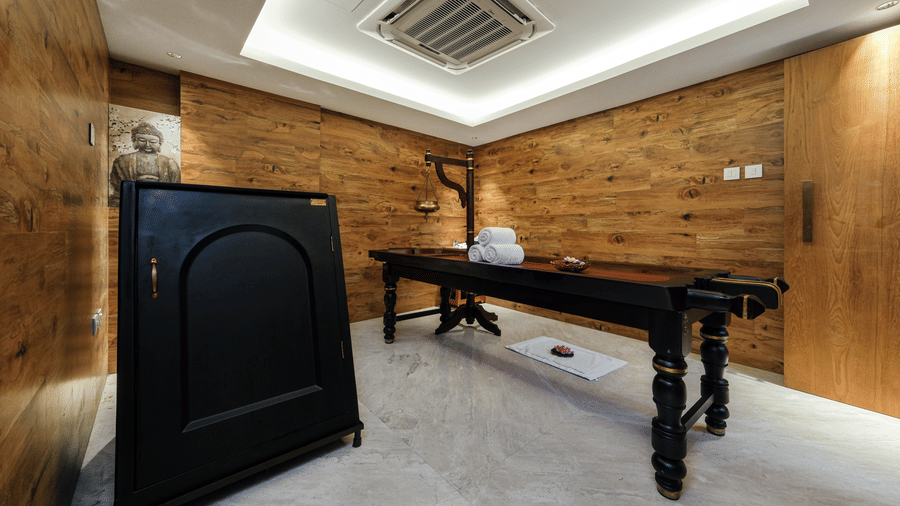 A wide shot of a wood-panelled treatment room at Tiara by MPS showcasing a traditional black wooden massage table and a steam cabinet in the corner.