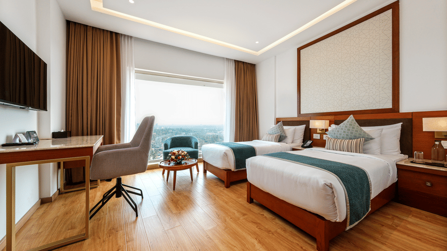 A twin bedroom featuring 2 beds, a study desk with a chair, and a large window presenting an outdoor view over trees at Tiara by MPS.