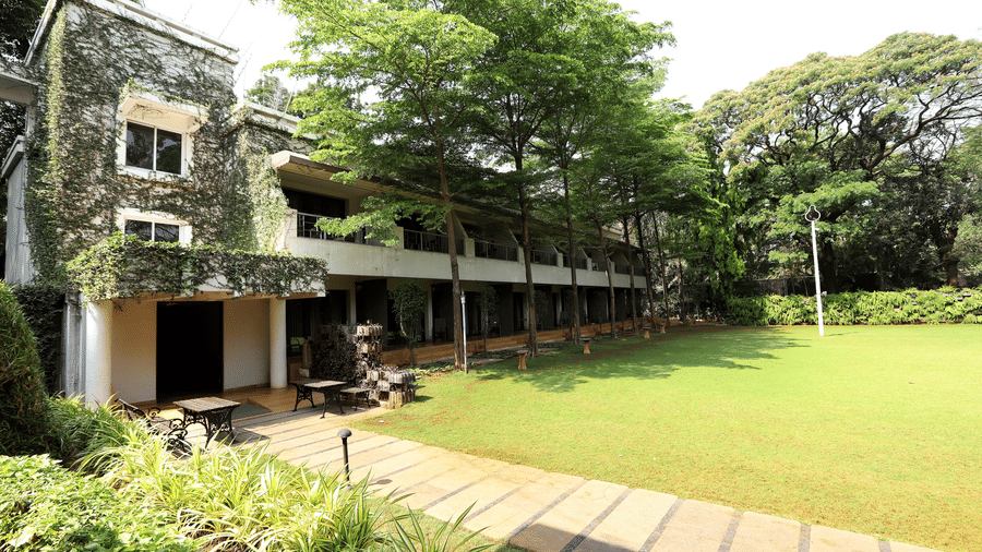 Exterior of a 2-storey stone building at Trance Sunderban Resort surrounded by tall green trees and a sunlit paved walkway.