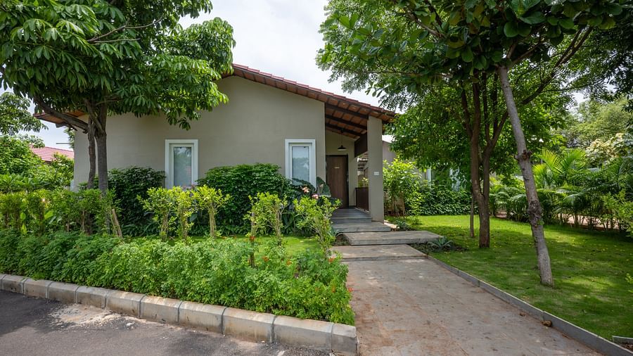 The front view of Ananta Garden Villa, a light-colored house with a red-tiled roof, surrounded by green trees and landscaping, with a pathway leading to the entrance.